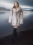 Woman wearing a fur coat standing by a body of water with mountains in the background