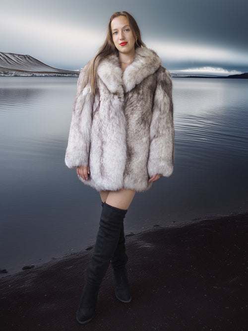 Woman wearing a fur coat standing by a body of water with mountains in the background