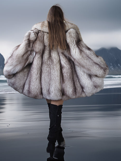 Person wearing a fur coat walking on a beach with mountains in the background