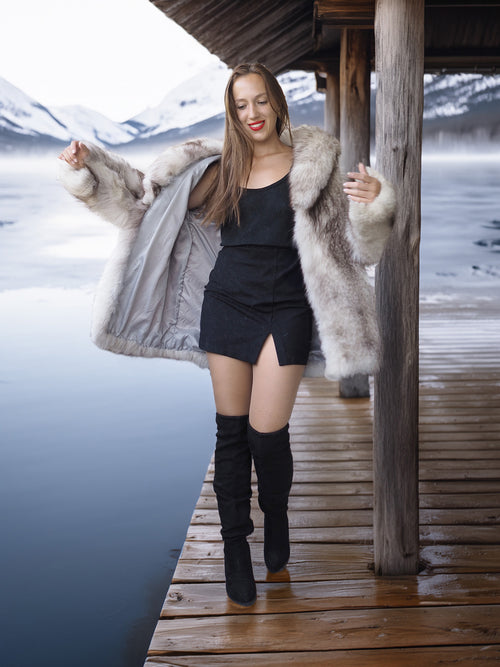 Woman in a fur coat and black outfit standing on a wooden deck by a snowy landscape.