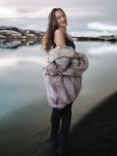 Woman wearing a fur coat standing by a body of water with a scenic background