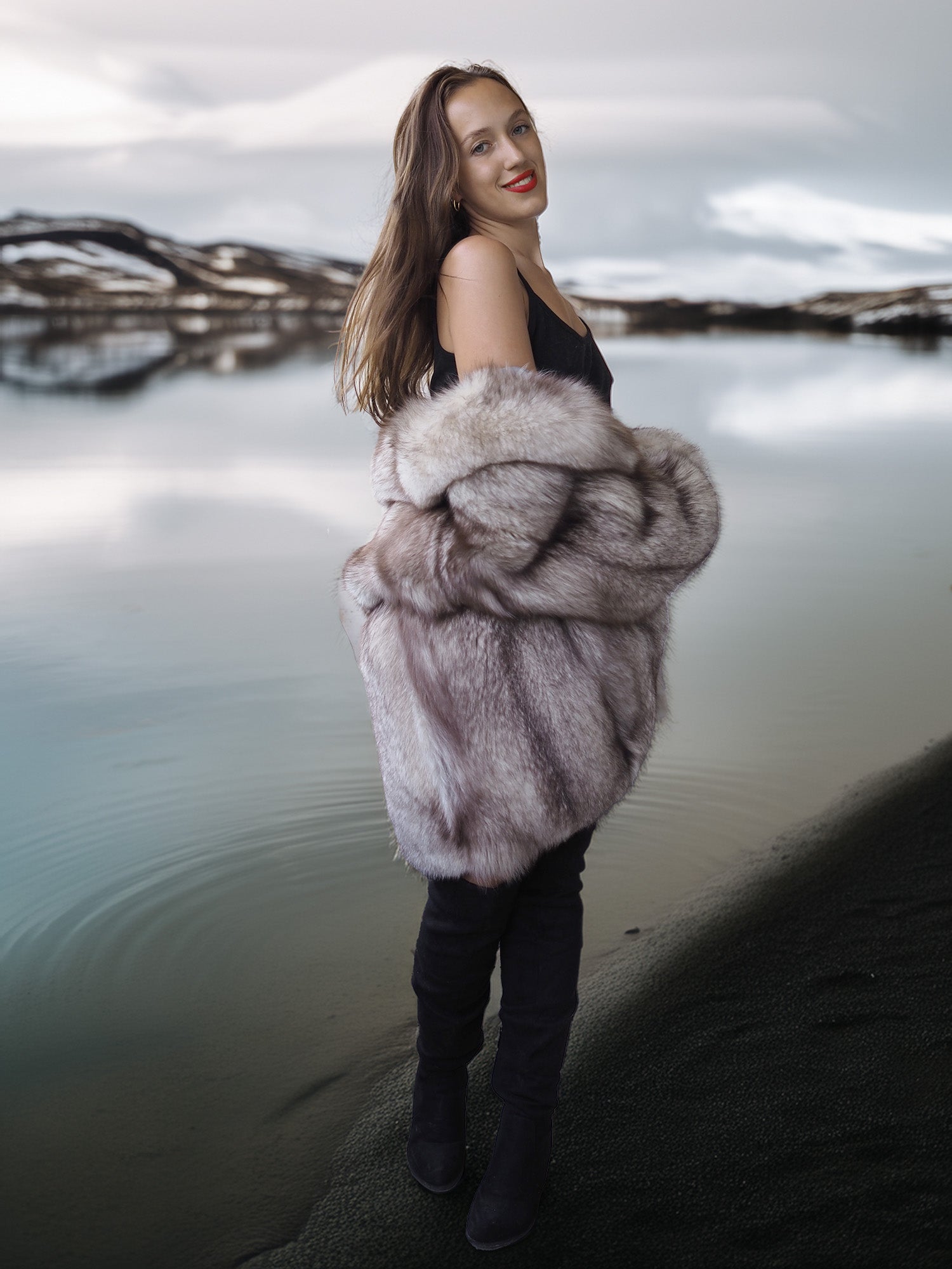 Woman wearing a fur coat standing by a body of water with a scenic background
