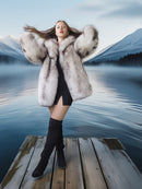 Woman in a fur coat and knee-high boots standing on a dock with a mountainous landscape in the background