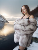 Woman in a fur coat standing by a lake with mountains in the background