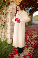 Woman in a long white fur coat standing on a path with red flower petals, stone wall, and archway in the background.