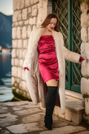 Woman in a pink dress and fur coat standing against a stone wall.