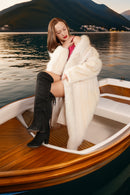 Woman in a white fur coat and knee-high boots on a boat with a mountainous background