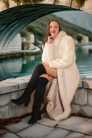 Woman in a white fur coat and black knee-high boots sitting by a body of water with a bridge in the background.