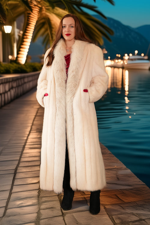 Woman in a long white fur coat standing by a waterfront at dusk.