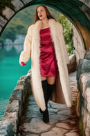 Woman in a red dress and white fur coat standing on a stone bridge over a body of water.