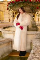 Woman in a white fur coat and red gloves standing by a fountain in an elegant outdoor setting.