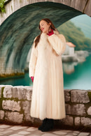 Woman in a long cream coat standing in front of a stone wall with a scenic background