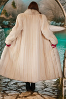 Person wearing a long beige fur coat standing in front of a scenic background.