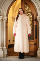 Woman wearing a long white fur coat standing in front of a wooden door with intricate design.