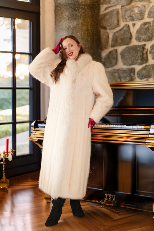 Woman in a long white fur coat standing in a room with a stone wall and piano.