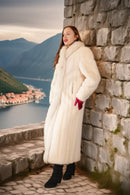Woman in a white fur coat standing against a stone wall with a scenic background