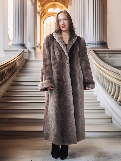 Woman wearing a long brown coat standing in an elegant interior setting with columns and stairs.