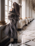 Woman in a long coat standing in a grand hallway with high ceilings and large windows.