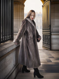 Woman in a long gray coat standing in an elegant interior setting.