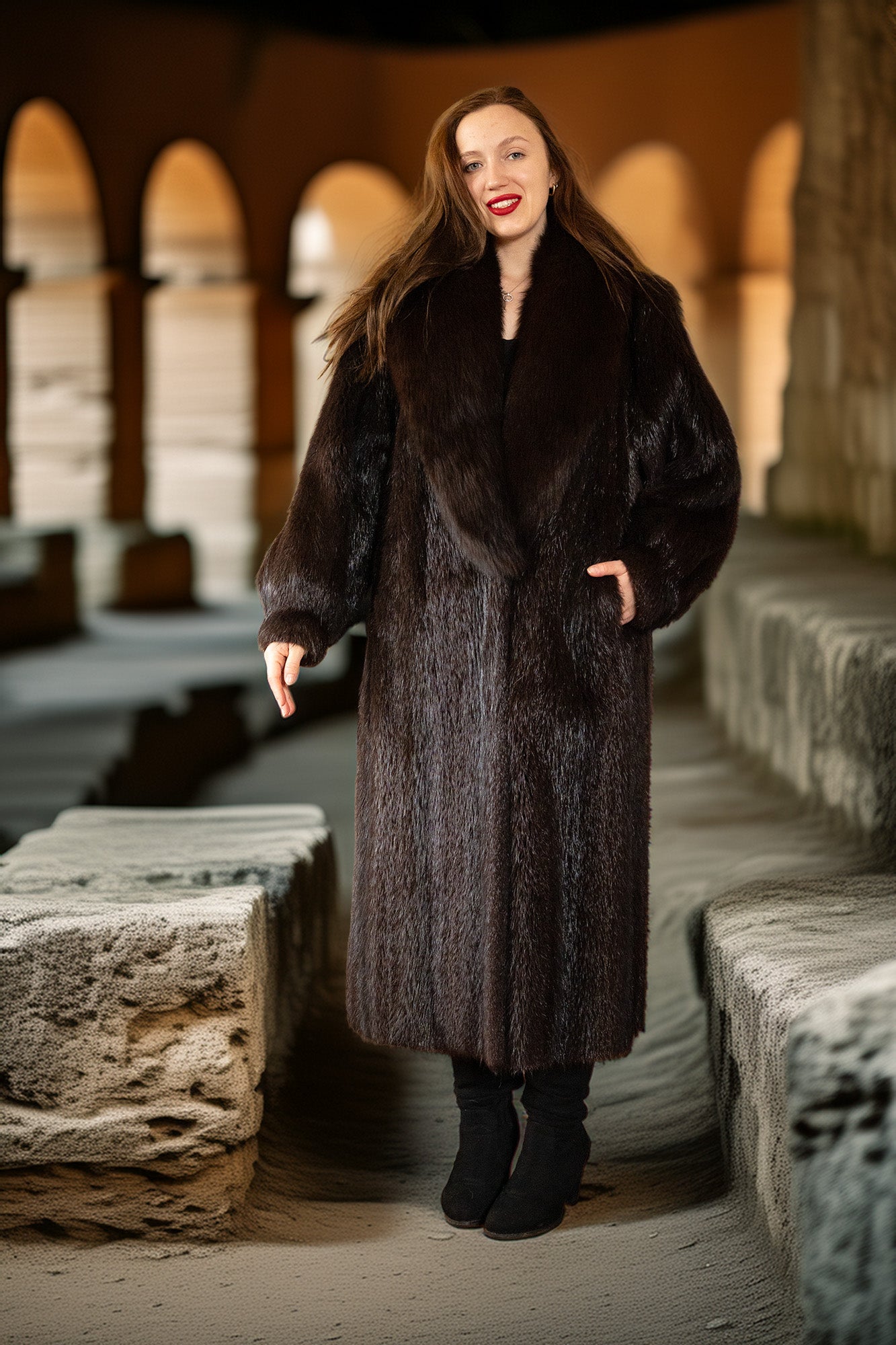 Woman wearing a long dark fur coat standing in an outdoor stone setting.