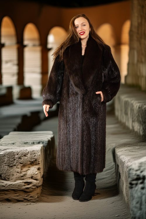 Woman wearing a long dark fur coat standing in an outdoor stone setting.