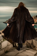 Person wearing a long dark coat standing on a stone path by a body of water with a cityscape in the background.
