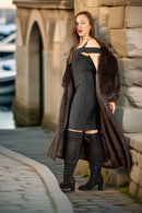 Woman in a long brown coat standing by a stone wall with a bridge and water in the background
