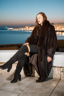 Woman in a fur coat and black boots sitting by a waterfront at night.