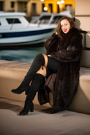 Woman in a fur coat sitting by a waterfront with boats in the background