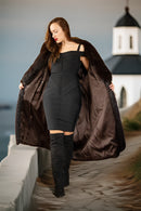 Woman in a black dress and brown coat standing on a stone wall with a scenic background