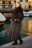 Woman in a fur coat standing by a waterfront with boats in the background