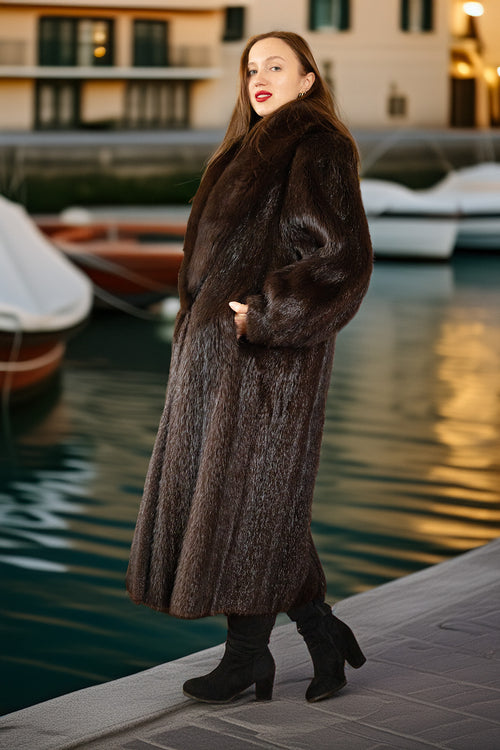 Woman in a fur coat standing by a waterfront with boats in the background