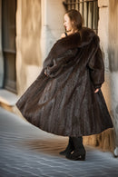 Woman wearing a long brown fur coat standing against a textured wall.