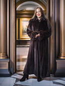 Woman in a long brown fur coat standing in an elegant interior setting.