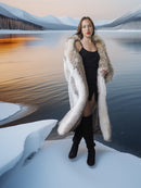 Woman in a long fur coat standing in a snowy landscape with a lake and mountains in the background.