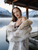 Woman in a fur coat standing by a lake with mountains in the background