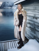 Woman in a fur coat standing on a dock by a body of water with mountains in the background