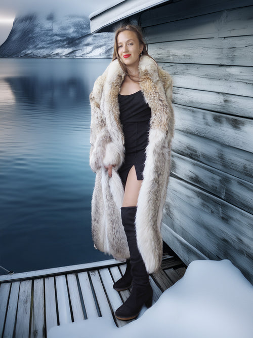 Woman in a fur coat standing on a dock by a body of water with mountains in the background