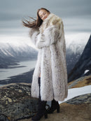 Person wearing a long fur coat standing on a mountainous landscape with snow and water.