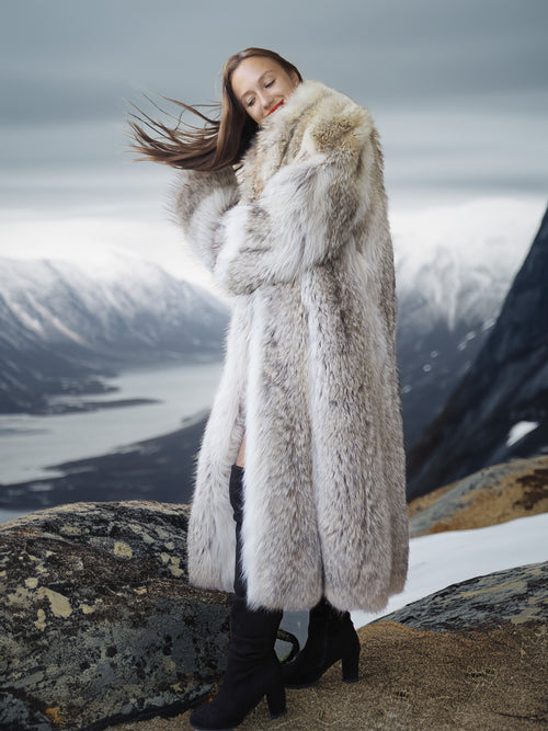 Person wearing a long fur coat standing on a mountainous landscape with snow and water.