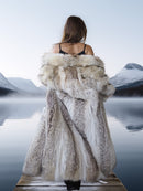 Person wearing a fur coat standing on a dock with a scenic lake and mountains in the background