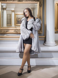 Woman in a gray fur coat standing in an elegant interior setting with framed pictures on the wall.