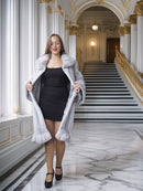 Woman in a gray fur coat standing in an elegant hallway with marble floors and gold accents.