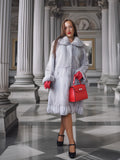 Woman in a gray fur coat and red gloves holding a red handbag in an elegant interior setting.