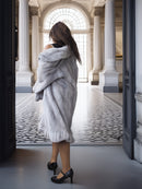 Woman in a fur coat standing in an elegant interior setting with columns and a patterned floor.