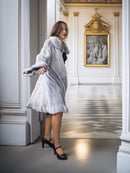 Woman in a gray fur coat standing in an elegant interior setting with classical art on the wall.