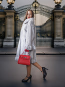 Woman in a fur coat holding a red handbag in front of an ornate gate.