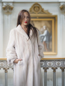 Woman wearing a white fur coat in an elegant interior setting with a painting in the background.