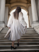 Person wearing a white fur coat walking up stone steps with classical architecture in the background