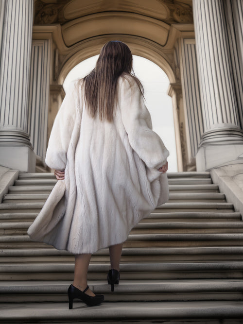 Person wearing a white fur coat walking up stone steps with classical architecture in the background